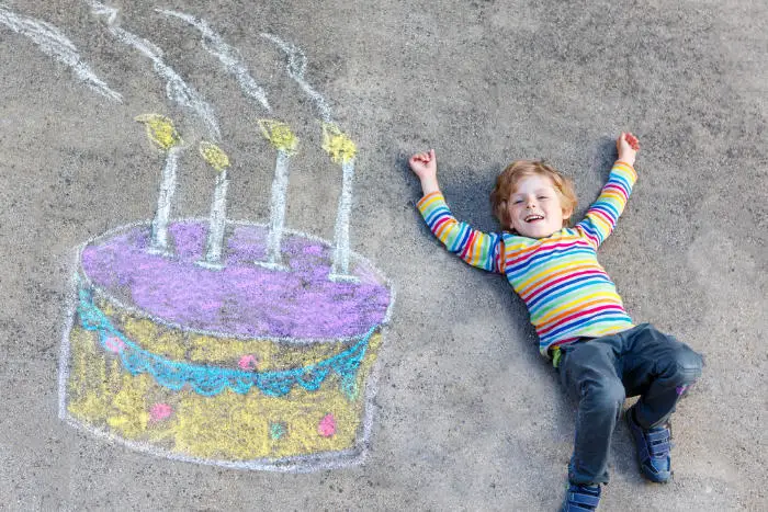 A sidewalk chalk drawing of a large birthday cake with a boy lying down beside it