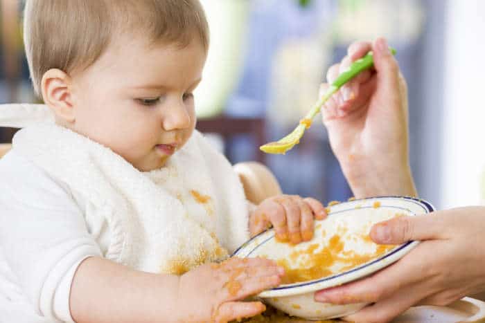 Baby boy being fed from a spoon but using his fingers and making a mess